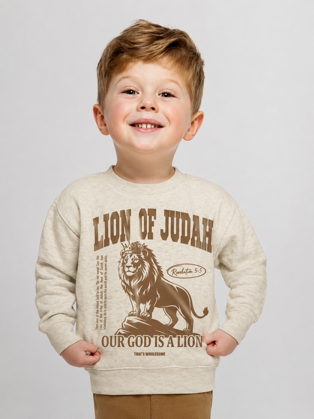 Toddler boy smiling, wearing a heather grey fleece crewneck with a brown graphic reading "Lion of Judah," an illustration of a crowned lion with "Revelation 5:5" in a small banner beside it, and "Our God is a Lion" beneath.