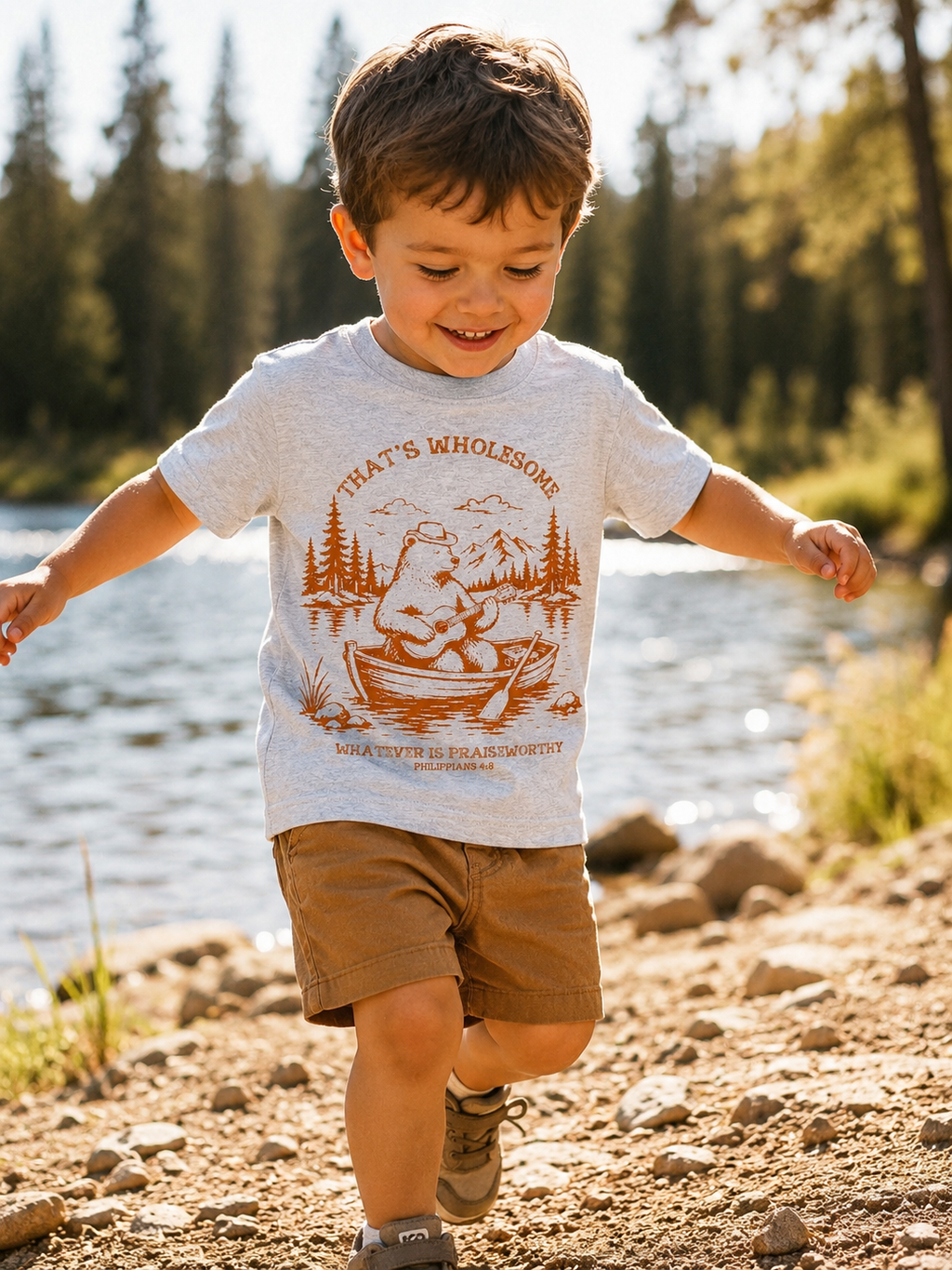 Toddler boy running along a rocky lakeshore wearing a heather gray short-sleeve t-shirt with a rust-colored graphic of a bear rowing a boat with a guitar, surrounded by pine trees and mountains, with the text "That's Wholesome" and "Whatever is Praiseworthy, Philippians 4:8."