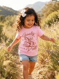 Toddler girl with a white bow running through a wildflower field wearing a pink short-sleeve t-shirt with a rust-colored graphic of a cowboy on a rearing horse, script text "Lovely" and "Y'all," and "Philippians 4:8" beneath the horse.