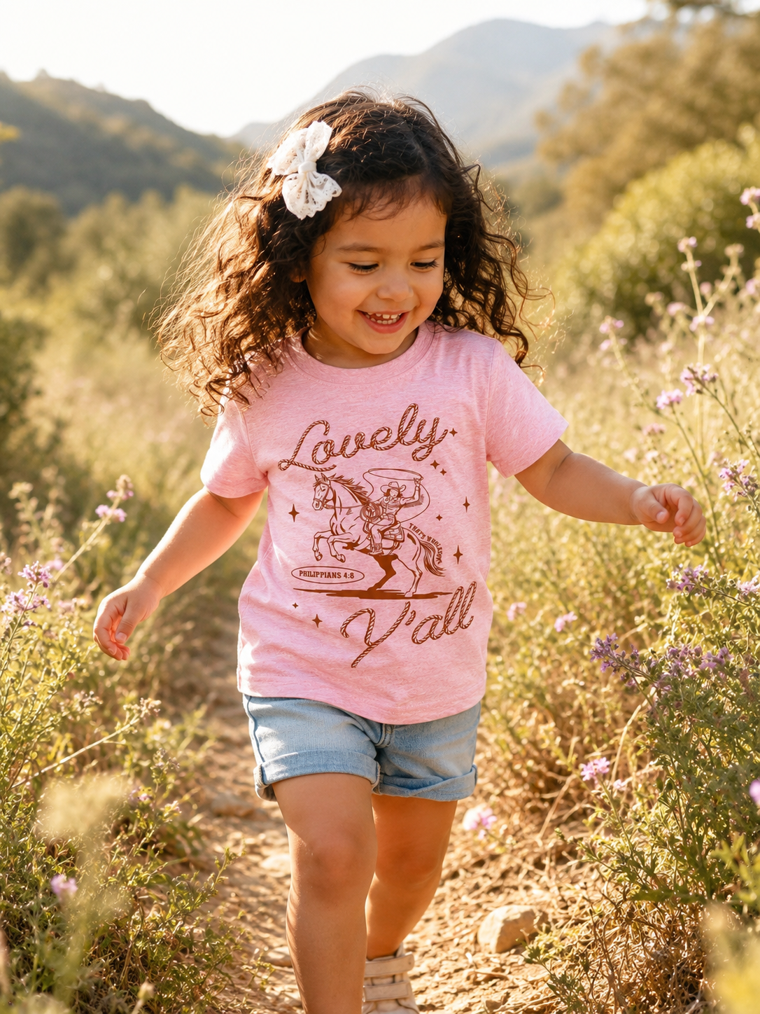 Toddler girl with a white bow running through a wildflower field wearing a pink short-sleeve t-shirt with a rust-colored graphic of a cowboy on a rearing horse, script text "Lovely" and "Y'all," and "Philippians 4:8" beneath the horse.