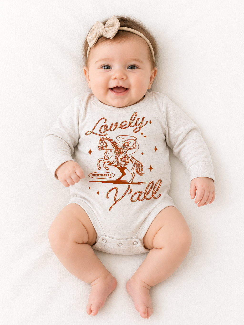 Smiling baby wearing a bow headband, lying on a white blanket in a beige long-sleeve onesie with a rust-colored graphic of a cowboy swinging a lasso while riding a rearing horse, rope-style script reading "Lovely" above and "Y'all" below, with "Philippians 4:8" and "That's Wholesome" in a small banner beneath the horse.