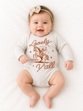 Smiling baby wearing a bow headband, lying on a white blanket in a beige long-sleeve onesie with a rust-colored graphic of a cowboy swinging a lasso while riding a rearing horse, rope-style script reading "Lovely" above and "Y'all" below, with "Philippians 4:8" and "That's Wholesome" in a small banner beneath the horse.