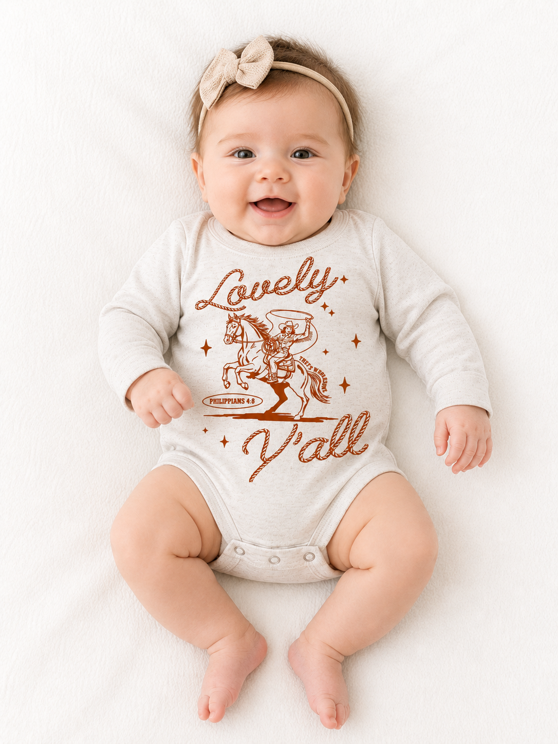 Smiling baby wearing a bow headband, lying on a white blanket in a beige long-sleeve onesie with a rust-colored graphic of a cowboy swinging a lasso while riding a rearing horse, rope-style script reading "Lovely" above and "Y'all" below, with "Philippians 4:8" and "That's Wholesome" in a small banner beneath the horse.
