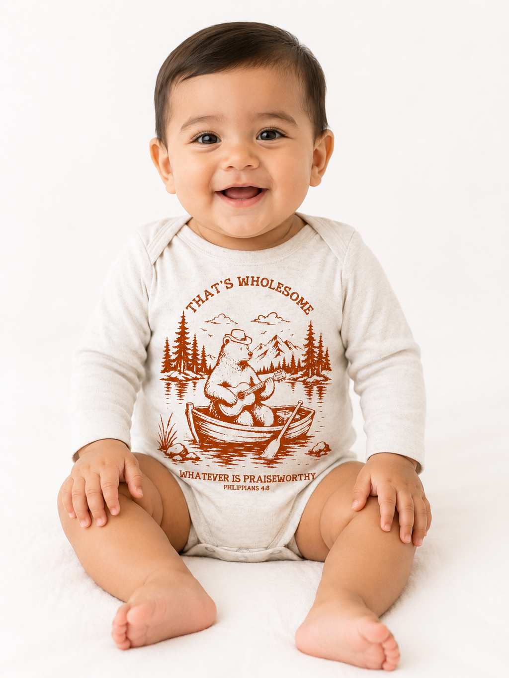 Smiling baby sitting on the floor wearing a beige long-sleeve baby onesie with a rust-colored graphic of a bear playing guitar in a rowboat, with the text "That's Wholesome" and "Whatever is Praiseworthy, Philippians 4:8".