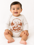 Smiling baby sitting on the floor wearing a beige long-sleeve baby onesie with a rust-colored graphic of a bear playing guitar in a rowboat, with the text "That's Wholesome" and "Whatever is Praiseworthy, Philippians 4:8".