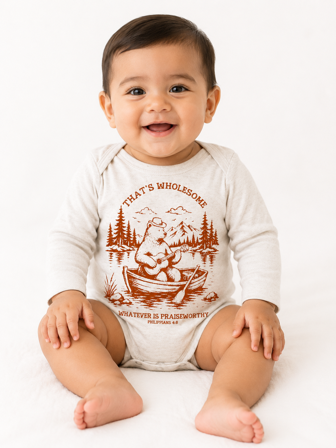 Smiling baby sitting on the floor wearing a beige long-sleeve baby onesie with a rust-colored graphic of a bear playing guitar in a rowboat, with the text "That's Wholesome" and "Whatever is Praiseworthy, Philippians 4:8".