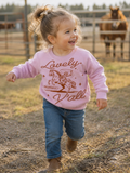 Toddler girl with a high ponytail running on a ranch, wearing a pink fleece crewneck with a rust-colored graphic of a cowboy swinging a lasso while riding a rearing horse, rope-style script reading "Lovely" above and "Y'all" below, with "Philippians 4:8" and "That's Wholesome" in a small banner beneath the horse.